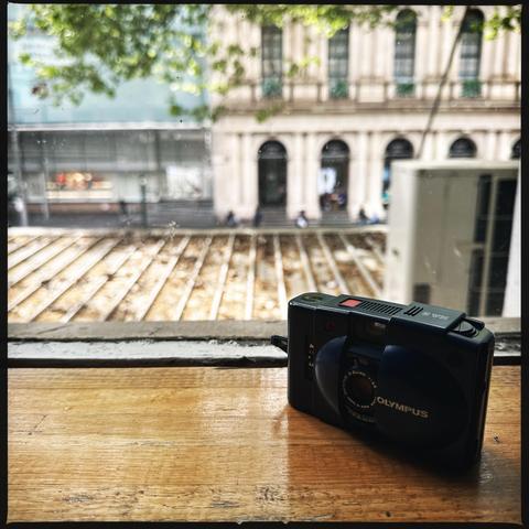 An Olympus XA2 film camera in the bottom right corner of the frame, on a windowsill with a first-floor view of a roof below us and an old building in the distance, out of focus. Some tree branches reach down from above with green leaves, also out of focus.