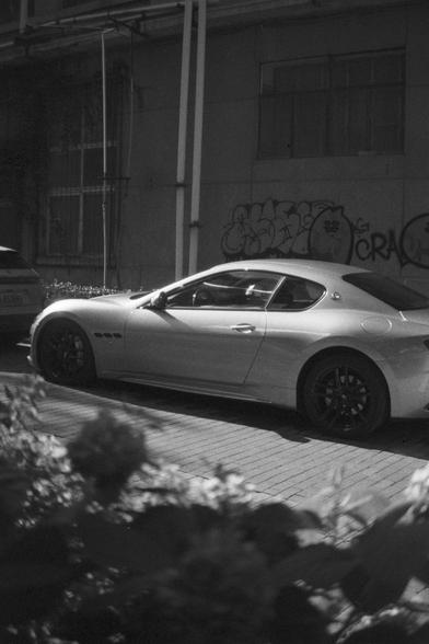 Ilford HP5 Plus 400 (FF)

English Alt Text:
A silver Maserati sports car is parked on a brick-paved street beside a wall covered in graffiti. The car has sleek curves, black alloy wheels, and a modern design. The photo is in black and white, giving it a timeless, cinematic feel. Blurred foliage in the foreground adds depth. Another vehicle is partially visible nearby. The contrast between the luxury car and gritty urban surroundings creates a visually compelling scene.

中文替代文字：
一辆银色玛莎拉蒂跑车停在砖铺街道旁，旁边的墙上有涂鸦。车辆线条流畅，配有黑色合金轮毂，设计现代。整张照片为黑白色调，营造出经典电影般的氛围。前景中有模糊的植物，增强了画面层次感。左侧还有另一辆车的部分。豪车与街头涂鸦的对比形成强烈视觉冲击。