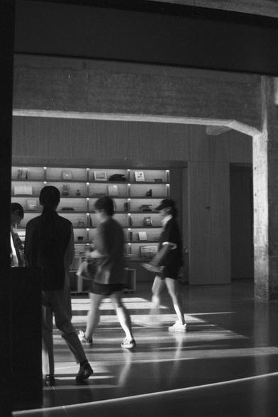 Ilford HP5 Plus 400 (FF)

English Alt Text:
A black-and-white photo of a modern indoor space with clean lines and minimalist design. Four people are walking or standing near a wall with built-in shelves filled with books and small objects. The floor is smooth and reflective, and the lighting casts soft shadows. Exposed concrete beams add an industrial touch. The scene feels like a gallery, museum, or bookstore, capturing quiet motion and thoughtful architecture.

中文替代文字：
这是一张黑白照片，展示一个现代室内空间，设计简约线条清晰。画面中有四人走动或站立在一面嵌入式书架前，书架上整齐摆放着书籍和小物件。地面光滑且有反光效果，灯光投下柔和阴影。裸露的混凝土梁增添工业风格。整体氛围像是画廊、博物馆或书店，展现静谧流动与建筑美感。