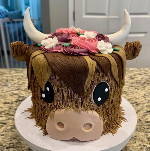A cake decorated to look like a cute highland cow with large black eyes, tan and brown fur, white horns, and a crown of pink and purple frosting flowers on its head.