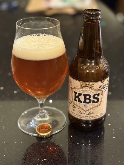 A snifter glass full of golden ale with a creamy white head. The short brown bottle is to the right with a white label and a gold accented “KBS” lettering in black