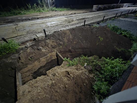 A night time snap of a partially dug up garden. They dug a trench where the tubes of the heat pump's source are running to check for leaks. There are piles of sand and weeds visible. In the background, there is a sandy road that's under construction, with the paving stones in the back, to be laid out next week.