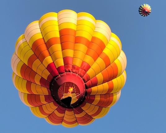 A hot air balloon flying overhead. It's shades of yellow, orange, and red with a triangular basket. The burner is firing. In the distance, in the top right corner and even farther up, is another balloon that appears to be rainbow-esque with dark vertical stripes.