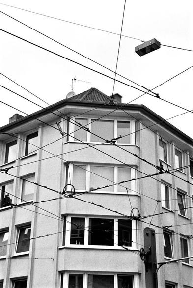 Looking up at overhead wires that crisscross over an intersection. In the background is an apartment building.
