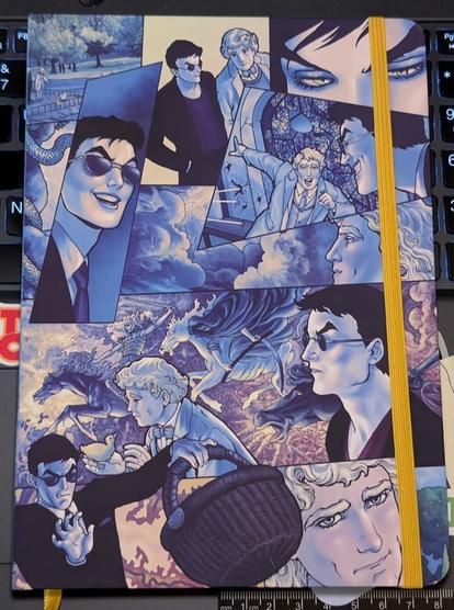 Photo of a notebook laying on a laptop keyboard. The notebook cover is filled with panels containing art from the Good Omens graphic novel. The vast majority of the art has a blue tone, there are some areas that have background colors which are more purple.

A yellow elastic strap runs vertically down the notebook holding it closed. A small piece of the yellow ribbon bookmark is visible coming out of the spine below the notebook.