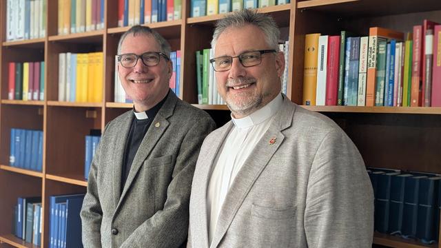 Zwei Herren mit graumeliertem Haar in Kollarhemd und Jackett stehen vor einem Bücherregal und lächeln freundlich in die Kamera. Beide tragen eine Brille. Der Bischof hat einen kurz geschnittenen Vollbart.