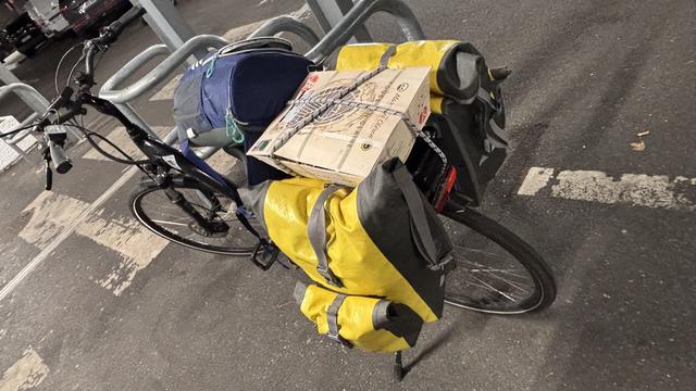 Un vélo noir aux sacoches gonflées de victuailles et avec une bourriche d’huîtres sur le porte bagage. Un sac à dos glacière plein est posé sur la selle.