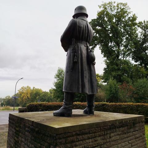 A larger-than-life sculpture of an armed Wehrmacht soldier from the First World War on a pedestal, overlooking a vast landscape. Photographed from behind.