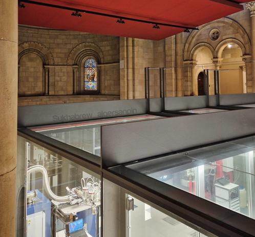 Interior of the Marenostrum supercomputer housed inside a deconsecrated neo-Romanesque chapel. Glass walls reveal the glowing machinery below, framed by stone arches and a stained-glass window above. The contrast between sacred architecture and advanced technology creates a serene, almost spiritual atmosphere.