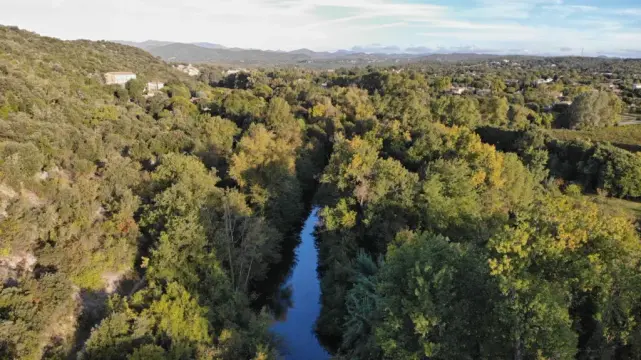 Vue aérienne d'un petit fleuve au milieu de la végétation. Plus loin, les maisons d'un village sous un ciel entre bleu et nuages d'altitude.