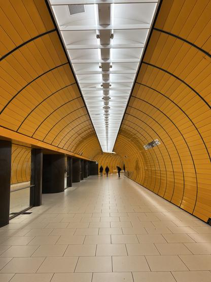 A modern subway tunnel with a continuous orange and white arching design. The floor is tiled, and a few silhouetted figures are walking in the distance, creating a sense of depth and perspective.