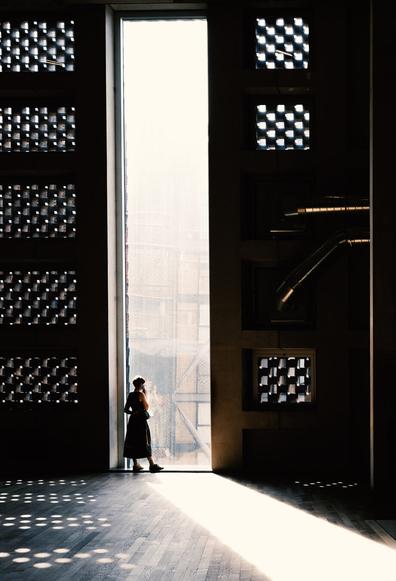 A silhouette of a lady on the background of a wall height window, she is looking out whilst holding the phone to her ear .the window is framed by walls with geometric patterns