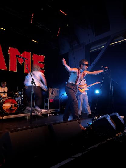 Shame on stage, vocalist with shades, no shirt, suspenders, arms wide, base was so busy that he’s just a blur in the air