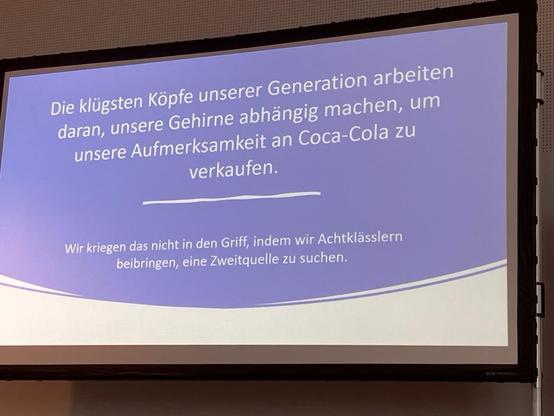 Eine Folie mit einem Beamer auf eine Leinwand projeziert. Auf dieser ist zu lesen: „Die klügsten Köpfe unserer Generation arbeiten daran, unsere Gehirne abhängig machen, um unsere Aufmerksamkeit an Coca-Cola zu verkaufen. Wir kriegen das nicht in den Griff, indem wir Achtklässlern beibringen, eine Zweitquelle zu suchen.“