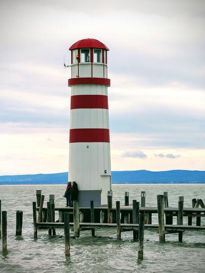 A tall, narrow lighthouse with alternating horizontal red and white stripes stands on a wooden pier over a lake. The lighthouse has a red dome and a small walkway near the top. Two people are standing near the base of the lighthouse on the pier. The water in the foreground is choppy, and the sky is overcast with heavy grey clouds. In the distance, a low shoreline or mountains are visible.