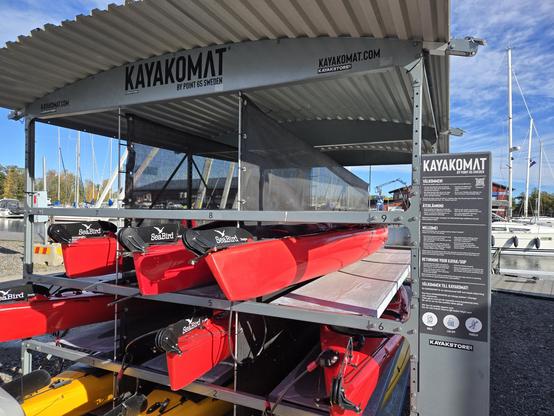 En öppen hyllan med ca sex röda havskajaker och två gula. Skylt "Kayakomat" och en informationsskylt om hur man hyr en kajak