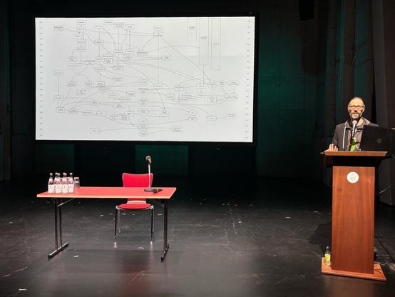 Ein Mann (Arne Vogelgesang) steht an einem Rednerpult auf einer Bühne und spricht in ein Mikrofon. Neben ihm hängt eine große Leinwand, auf der ein komplexes Diagramm mit Linien und Begriffen zur Entwicklung digitaler Technologien / XR zu sehen ist. Vor der Bühne steht ein roter Tisch mit einem weiteren Mikrofon, einem roten Stuhl und mehreren Wasserflaschen. Die Szene ist klar ausgeleuchtet, der Hintergrund bleibt dunkel und nüchtern – Betonwände, Theaterlicht, Konzentration auf Inhalt und Person. Die Atmosphäre wirkt sachlich, intellektuell und leicht theatral.