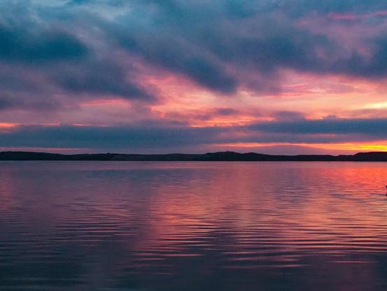 The image depicts a wide, calm body of water under a vibrant, colorful sky at dusk or dawn. The lower half of the image is dominated by the water’s surface, which is rippled with small waves, creating a shimmering, reflective texture. Above the water, a dark, silhouetted landmass stretches horizontally across the middle ground, appearing as a flat, unbroken shape. The sky is a gradient of colors, primarily shades of pink and orange, with deeper purple hues towards the top of the frame. The overall effect is one of tranquility and natural beauty, with a strong emphasis on color and reflection.