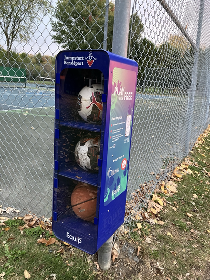 A vertical case holding three balls, two soccer balls and basketball beside a tennis court in a park in Ottawa
