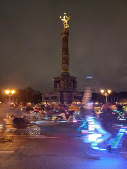 Ein Fotos vom großen Stern mit Goldelse. Die Goldelse ist zentral im Foto von nächtlichem Himmel. Davor fahren menschen auf Fahrrädern. Die Menschen und rader sind verschwommen, an den Silhoutten der Räder leuchten Lichter in blau, rot und gelb.