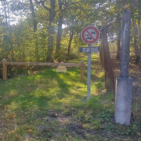 Photographie d'un chemin en forêt. Un panneau fixe indique que c'est un chemin rural. Un panneau temporaire jaune indique "Chasse en cours, tire à balles. Passage déconseillé, ensemble soyons vigilants"
Le chemin borde une clôture grillagée.