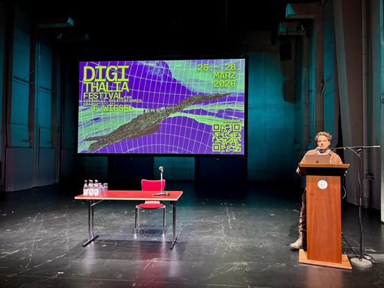 Jost von Harleßem     steht auf einer Bühne am Pult und spricht vor einer großen, bunt leuchtenden Projektion. Auf der Leinwand steht: „DIGITHALIA – Festival für virtuelle Theaterformen, Graz, 25.–28. März 2026“. Darunter der Künstlername Flink Wiesel und eine QR-Code-Grafik. Der Raum ist dunkel, die Bühne minimalistisch eingerichtet mit rotem Tisch, Stuhl, Mikrofon und Wasserflaschen.