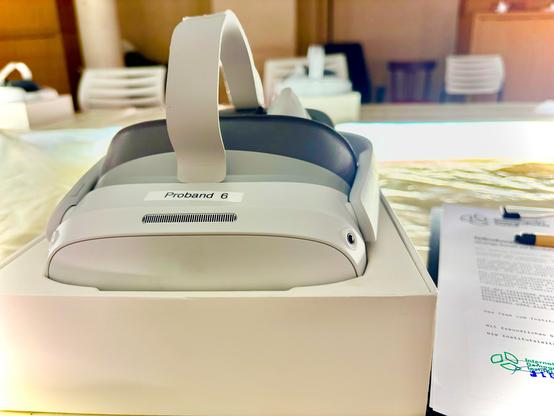 Eine Virtual-Reality-Brille liegt in einer weißen Box auf einem Tisch. Auf der Vorderseite klebt ein Etikett mit der Aufschrift „Proband 6“. Im Hintergrund sind weitere Geräte, Stühle und ein Formular mit dem Logo des „Instituts für …“ (unscharf erkennbar) zu sehen. Das Bild vermittelt eine ruhige, experimentelle Atmosphäre – wie in einem Forschungslabor oder einer Testumgebung für VR-Anwendungen.