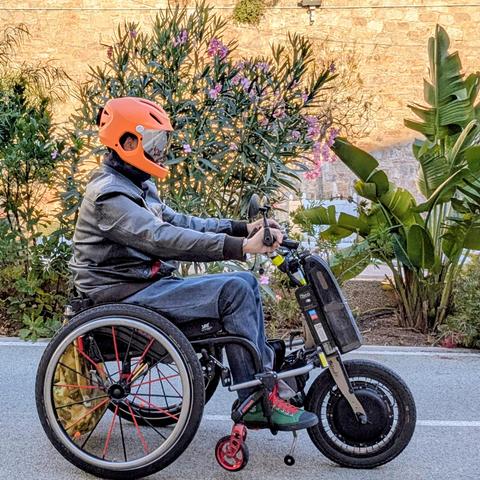 Photo de moi, en fauteuil roulant au volant de mon handbike électrique, de côté, je roule vers la droite.
Je porte un casque intégral orange.
Derrière moi, des feuillages.