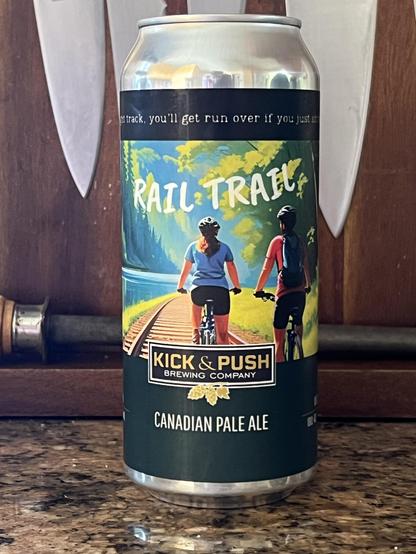 A can of beer on a kitchen counter. The label reads “Kick & Push Brewing Company” and “Rail Trail Canadian Pale Ale”. There’s a painterly illustration of two people cycling away from the viewer, riding on a train track in a scenic rural area.