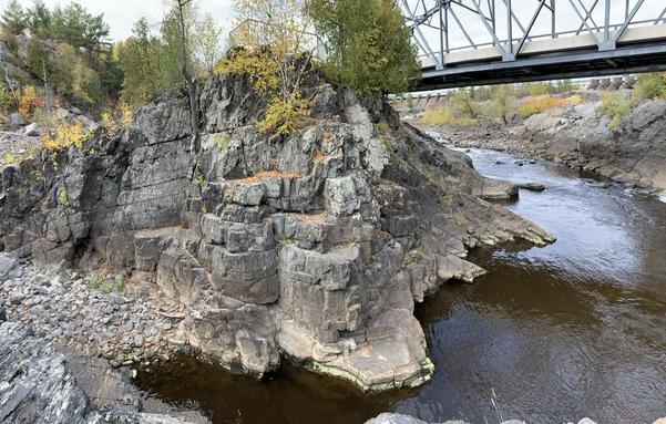 Outcrop along a riverbank showing a gray fine grained rock. Bedding planes dip in opposite directions on either side. Part of a bridge can be seen crossing the river.