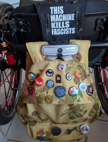 Photo de l'arrière de mon fauteuil roulant. Sur le dossier, un patch "This Machine Kills Fascists" et un sac à dos jaune tournesol de Van Gogh, dont le dessus et la poche devant sont recouvert de Badges et de pin's.
Firefox, no border only People, il n'y a pas d'étrangers sur cette terre, LDH, action antifasciste anarchiste, drapeau palestinien, légalisez le cannabis, handie queer, etc...
Il y a aussi un porte clef Daffy Duck lego accroché au dossier du fauteuil.