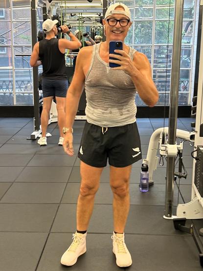 Me, taking a selfie in a gym, wearing a sleeveless, wet gray shirt and black shorts. They are smiling and holding a phone. In the background, another individual is using gym equipment and drinking from a bottle. The gym has large windows.
