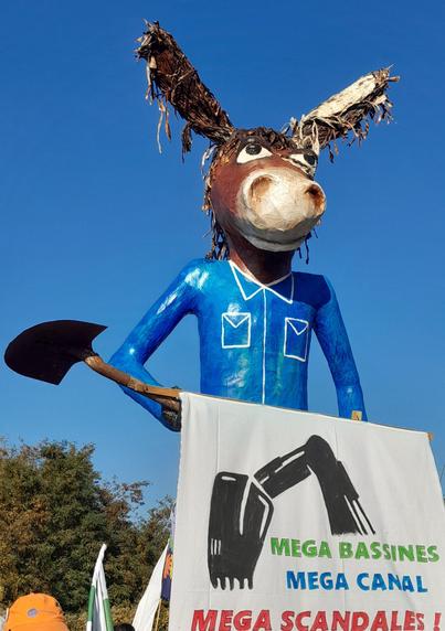 Un géant à tête d’âne vêtu d’un costume bleu, tenant un pelle dans les mains derrière une pancarte :
"MÉGA BASSINES
MÉGA CANAL
MEGA SCANDALES"