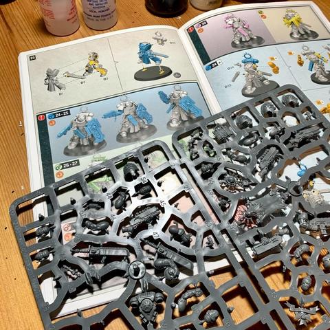 Pages from a Warhammer 40k instruction booklet showing how to construct models. On top of the booklet is a sprue of model pieces.