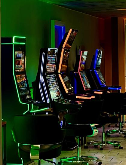 Blick in eine Spielhalle mit fünf bunt leuchtenden Glücksspielautomaten, davor jeweils ein schwarzer Drehstuhl mit Chromgestell
