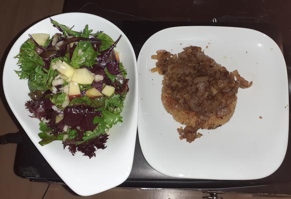 Es ist Links eine weiße Keramikschüssel (in Form eines umgedrehten Bauhelmes) gefüllt mit Salat sowie Rechts ein weißer quadratischer Keramikteller mit dem Haferschnitzel und den Zwiebeln auf dem Schnitzel zu sehen. Beides steht auf einem dunkelbraunen Couchtisch, fotografiert wurde von Oben.