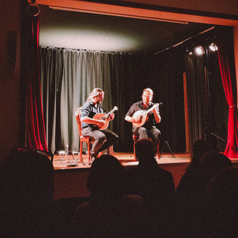 Two seated musicians, both bearded and long-haired on a cozy lit stage, each playing a small mandolin-like plucked instrument called waldzither.