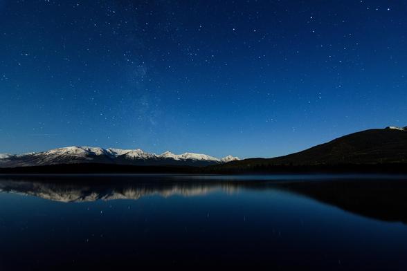 The blue, starry night sky, striped by the vertical rise of the Milky Way, rises above a snow-capped mountain range, sloping gently up from left, to a range, then back down again just right of centre and back up again to a taller mountain. Below, a still, deep lake reflects back the sky and mountains and flanking forest as in a mirror.