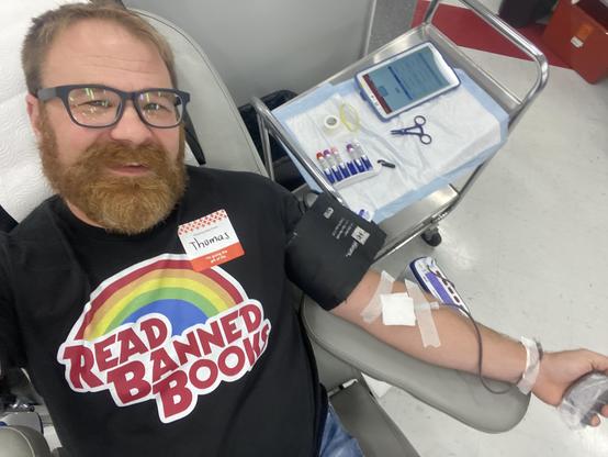 A bearded man in glasses is donating blood while wearing a T-shirt that says read band books under a rainbow reminiscent of the reading rainbow emblem