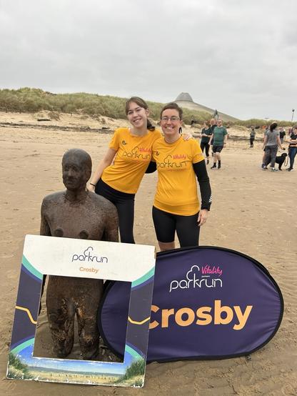 Emily and a teenage girl wearing apricot Parkrun tshirt standing behind a sign that says “Crosby”.