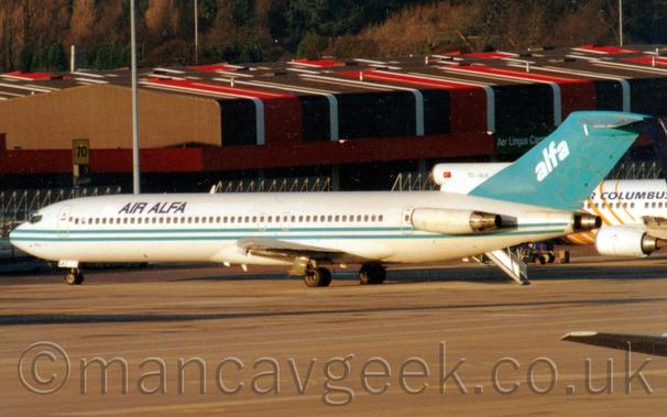 Side view of a 3 engined jet airliner with the engines mounted on the rear fuselage, parked facing to the left and slightly away from the camera.
The plane is mostly white, with a pair of thin, pale blue stripes running along the body just under the passenger cabin windows, and black "Air Alfa" titles on the upper forward fuselage.
The tail is the same blue, with diagonal, white, lower-case test "alfa" in the middle.
The centre engine intake is white, and has a small Turkish flag at the front, and the black registration "TC-ALK".
Concrete apron fills the foreground, with the wingtip of another plane just peeking into frame at the bottom right.
The forward fuselage of another jet airliner peeks into frame on the right of the background, with brown, white, and red cargo sheds stretching across the frame beyond that.
Trees in the distance complete the image.