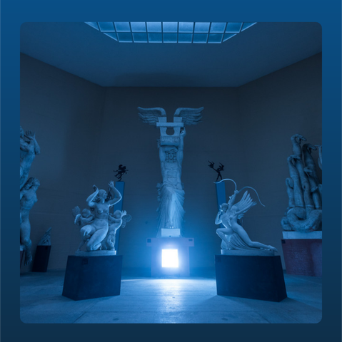 A dimly lit grand room with classical statues arranged on pedestals. At the far wall is a large central statue resembling a winged figure or angel, flanked by smaller sculptural works on either side. Light source: an intense, glowing square panel at floor level behind the statues illuminates the room with a cold blue glow. Overhead, a rectangular skylight is visible, casting pale light down. The overall color tone is blue-gray, giving the scene a solemn, eerie, almost ritualistic atmosphere.