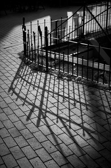 A black and white image showcasing the shadows of a curved metal fence on a textured brick surface, creating a striking interplay of light and shadow.