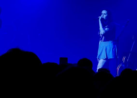 The photo captures a singer performing on stage under intense blue lighting. The performer stands slightly to the right, holding a microphone close to their mouth, mid-song. They wear a dark T-shirt and a short, light-coloured skirt. Their expression is focused, illuminated by the cool blue glow that fills the entire scene. Behind them, faint outlines of guitars and stage equipment are visible, while the foreground shows the dark silhouettes of the audience watching intently. The image conveys the quiet intensity of a live concert — a moment suspended between light, sound, and emotion, wrapped in an almost dreamlike blue haze.