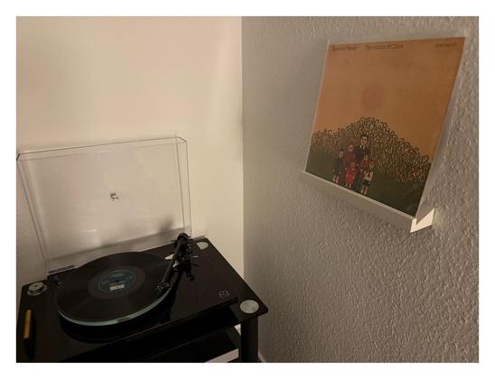 Auf einem schwarzen Plattenspieler läuft eine schwarze Schallplatte. Rechts an der Wand ist ein kleines weißes Regalbrett, darauf steht das Cover. Ein gemaltes Bild, eine sechsköpfige Familie steht vor einem großen Gebüsch. Darüber ist gelb-orangefarbener Himmel und die Sonne. Oben stehen Künstler und Titel.