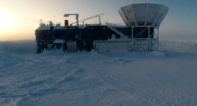A still from a video of someone walking from the Amundsen-Scott South Pole Station to the Dark Sector Laboratory. The image shows the moment when the person looks over at the Martin A. Pomerantz Observatory. In the desolate snow-covered expanse, is a 3-story asymmetrical structure with a medley of metal walls, structural lattice work, and jelly bean shaped windows. There's all kinds of vents, cranes and hoists on the roof. To the right side is what looks to be a large, immovable dish pointing up. This is part of the array of BICEP3 microwave telescopes observing inflation in the Cosmic Microwave Background.