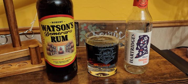 A glass of rum and cola between a bottle of Watson's Demerara Rum with a yellow label and a small glass bottle of Hartridges Cola with a white label sitting on a wooden worktop.