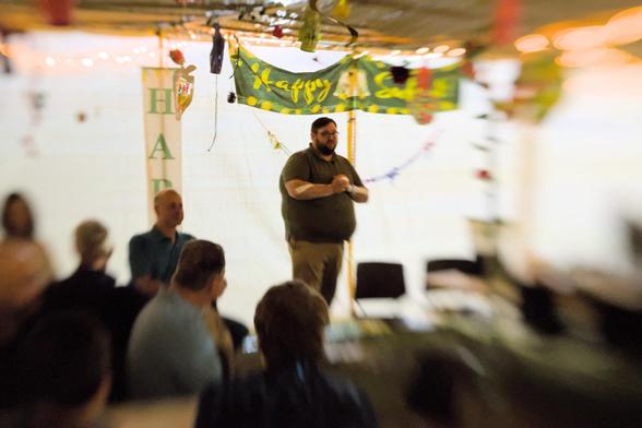 A modern rabbi standing in a sukkah. The back wall is backlit bright white behind him. The center of the photo is sharp. The edges get artistically blurry. There is a bright green sign in the background with yellow letters reading "Happy Sukkot"