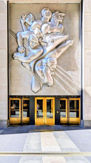 A view of the entrance to 50 Rockefeller Plaza in New York City. A large, stylized silver relief sculpture is mounted above the golden revolving doors.
