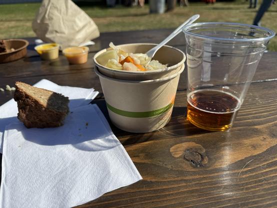 A picnic table bathed in sunlight holds a slice of bread, a bowl of vegetable and pork strew, and a nearly empty plastic cup of amber beer.